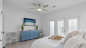 A bedroom with a bed, a ceiling fan, a TV, a blue cabinet, a chair, and a window with blinds. - Enjoy a personal beach experience! Expansive gulf side deck! - Dauphin Charm (Dauphin Island)