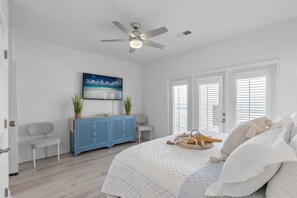 Un dormitorio con cama, ventilador de techo, televisión, un mueble azul, una silla y ventana con persianas.