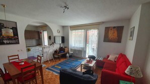 Un salon avec un canapé rouge, un fauteuil noir et un coin repas comprenant une table et des chaises en bois. Au fond, on aperçoit une cuisine avec une fenêtre et une horloge au mur.
