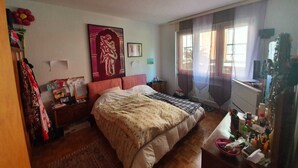 Une chambre à coucher avec un grand lit, une table de chevet, une commode, une fenêtre avec des rideaux et un mur orné d’un tableau encadré.