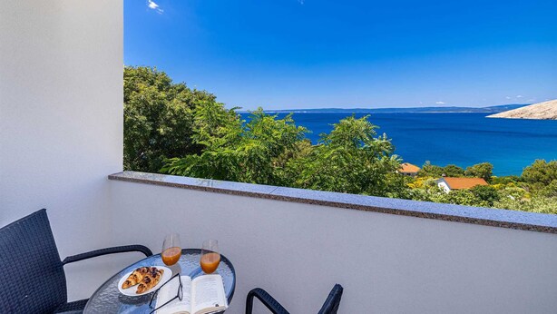 A balcony with a table set for two, overlooking a coastal view with trees and the sea. - Apartment STARA BAŠKA near the sea (Stara Baška)