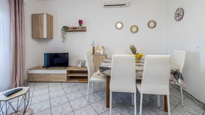 A modern living room with a dining area, featuring a TV, a wooden cabinet, a small shelf with plants, and a wall-mounted air conditioner. - Apartment STARA BAŠKA near the sea (Stara Baška)