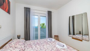 A hotel room with a bed, a small stool, a nightstand, a lamp, a large window with curtains, and a mirror on the wall. - Apartment STARA BAŠKA near the sea (Stara Baška)