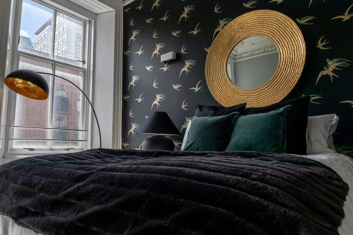 Une chambre avec un grand lit, un miroir rond et une fenêtre donnant sur des immeubles.
