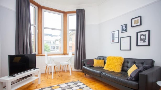 A modern living room with a black leather sofa, a white TV stand, a dining table with chairs, and a large window with curtains. - Beautiful Flat in Great West end Location (Glasgow City)