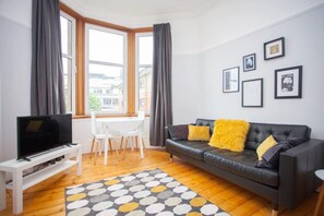 Una sala moderna con un sofá de cuero negro, un mueble de TV blanco, una mesa de comedor con sillas y un ventanal con cortinas.