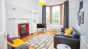 A living room with a fireplace, a TV, a sofa, a dining table, and a large window with curtains. - Beautiful Flat in Great West end Location (Glasgow City)