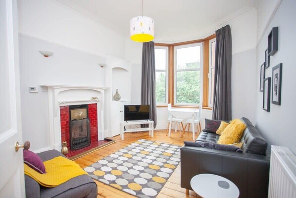 Una sala de estar con chimenea, televisor, sofá, mesa de comedor y un amplio ventanal con cortinas.