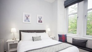 A neatly arranged hotel room with a bed, bedside tables, a window with a view, and two wall-mounted pictures. - Beautiful Flat in Great West end Location (Glasgow City)