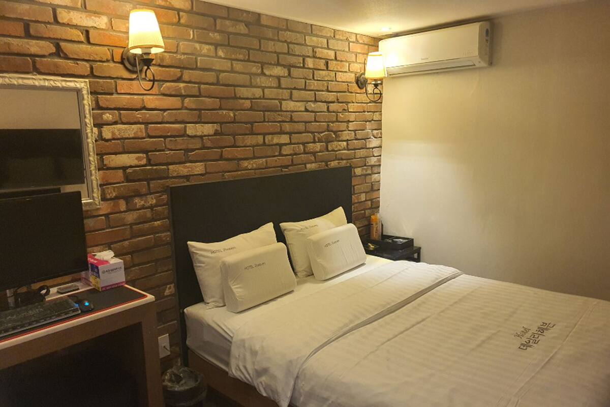 A hotel room with a brick wall, a bed with white bedding, a desk with a computer, and a trash can.