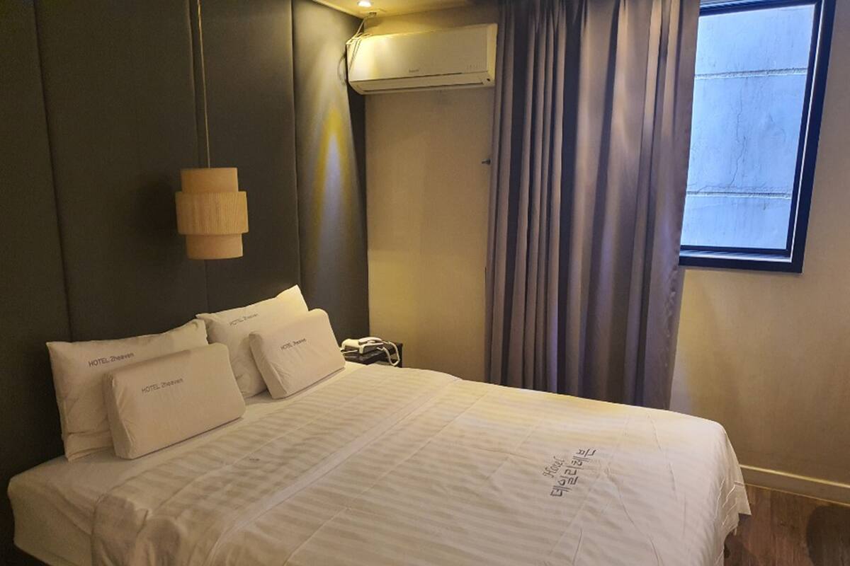 A hotel room with a bed, pillows, a bedside table, an air conditioning unit, and a window with curtains.