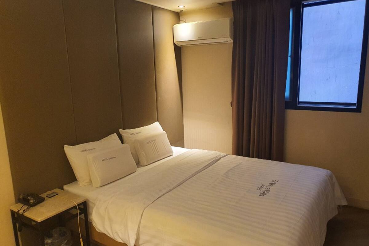 A hotel room with a bed, pillows, a nightstand, a telephone, and an air conditioning unit.