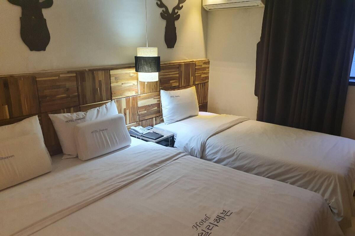 A hotel room with two beds, each with white bedding and pillows labeled "HOTEL ZOOZAVEN". There is a wooden headboard, a wall-mounted lamp, and a deer's head decoration.
