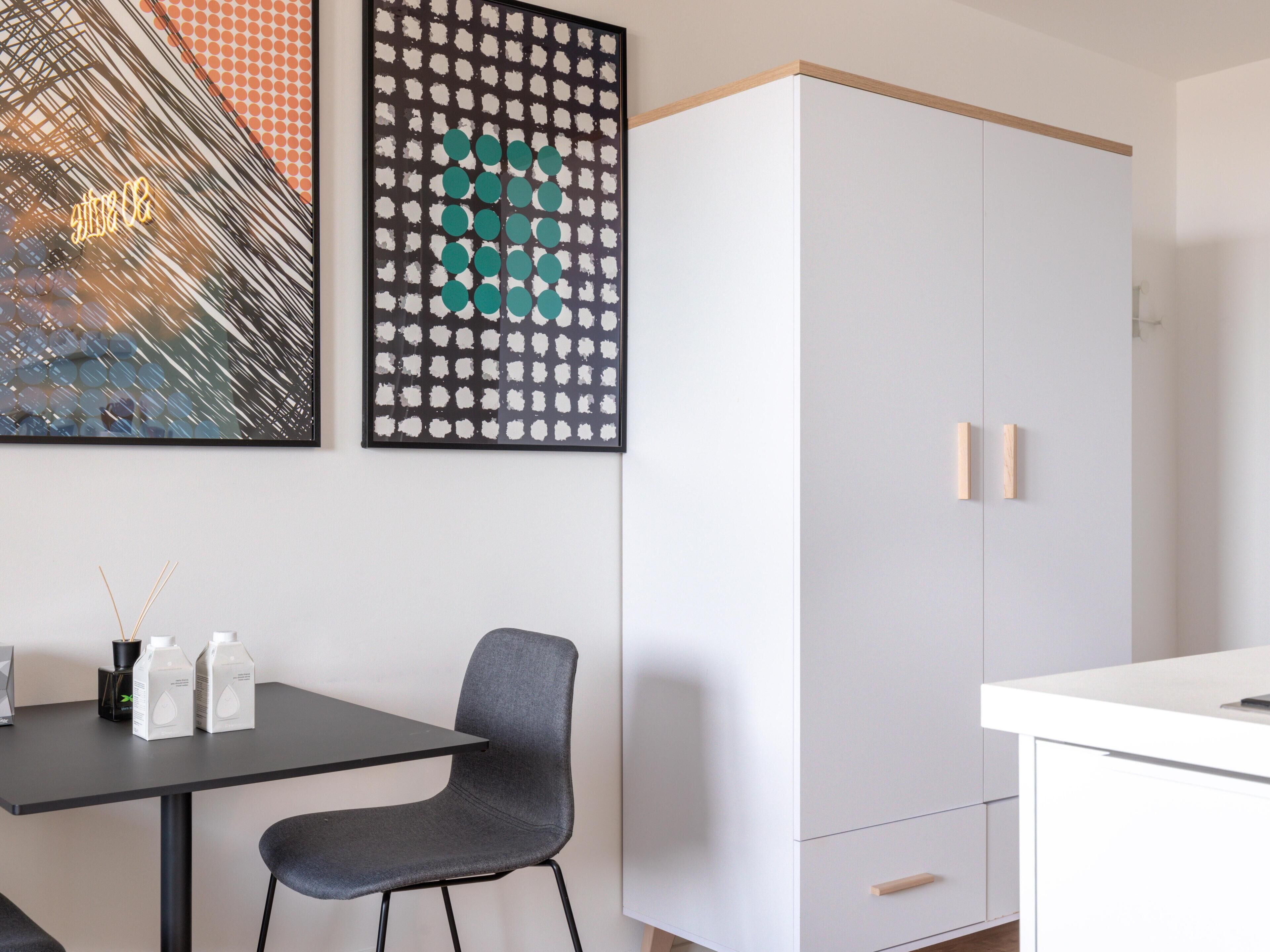Una habitación moderna con un armario blanco, una silla gris y dos obras de arte enmarcadas en la pared.