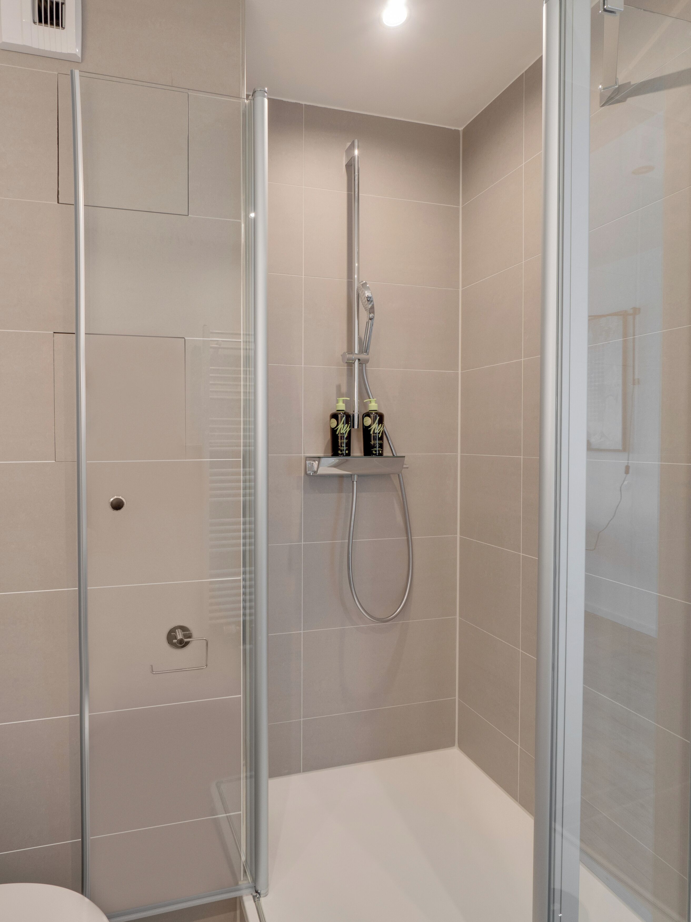 A modern shower with a glass enclosure, a handheld showerhead, and two bottles of shampoo on a shelf.
