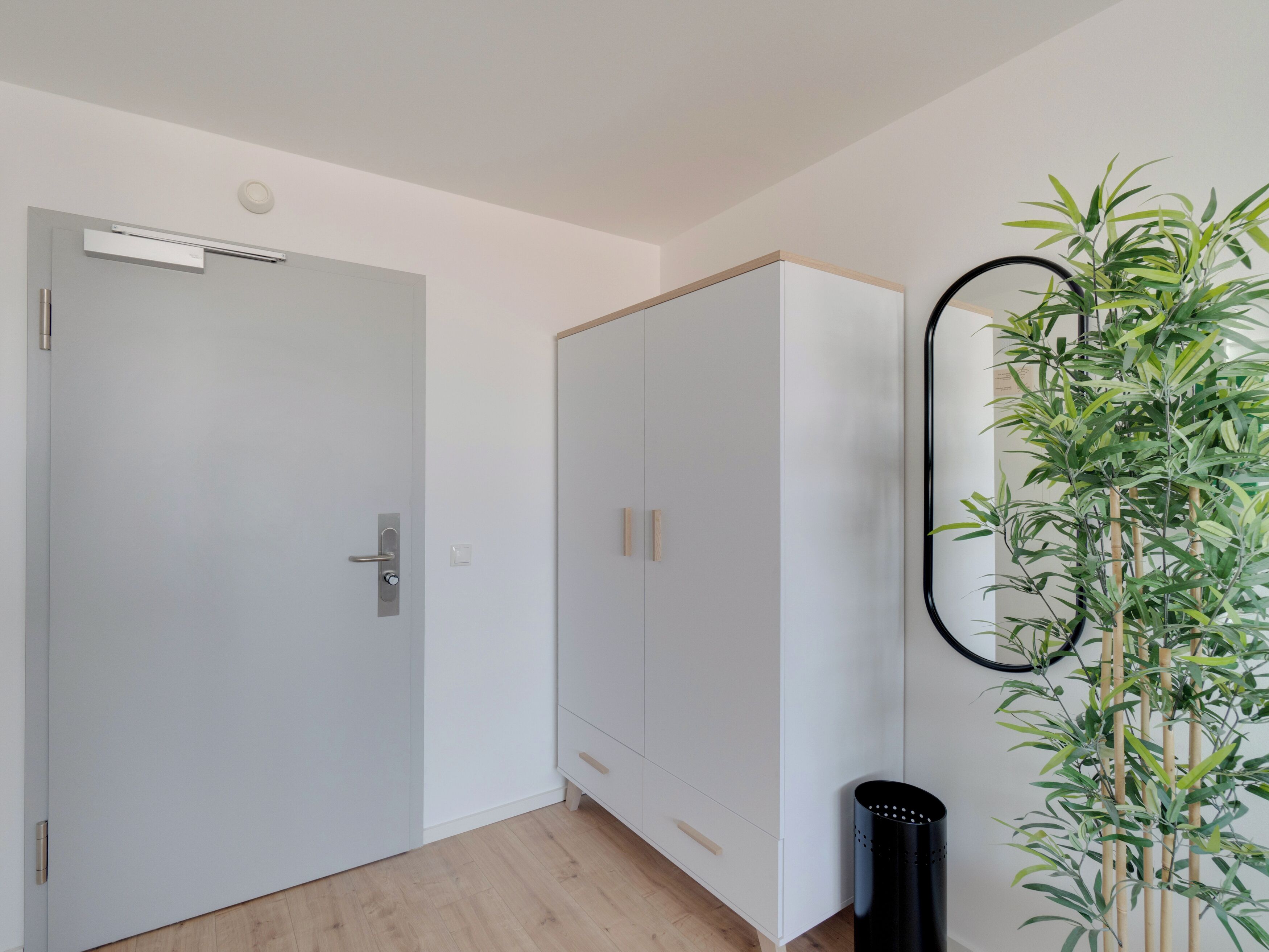 A room with a white wardrobe, a door with a handle, a light switch, and a black trash can.
