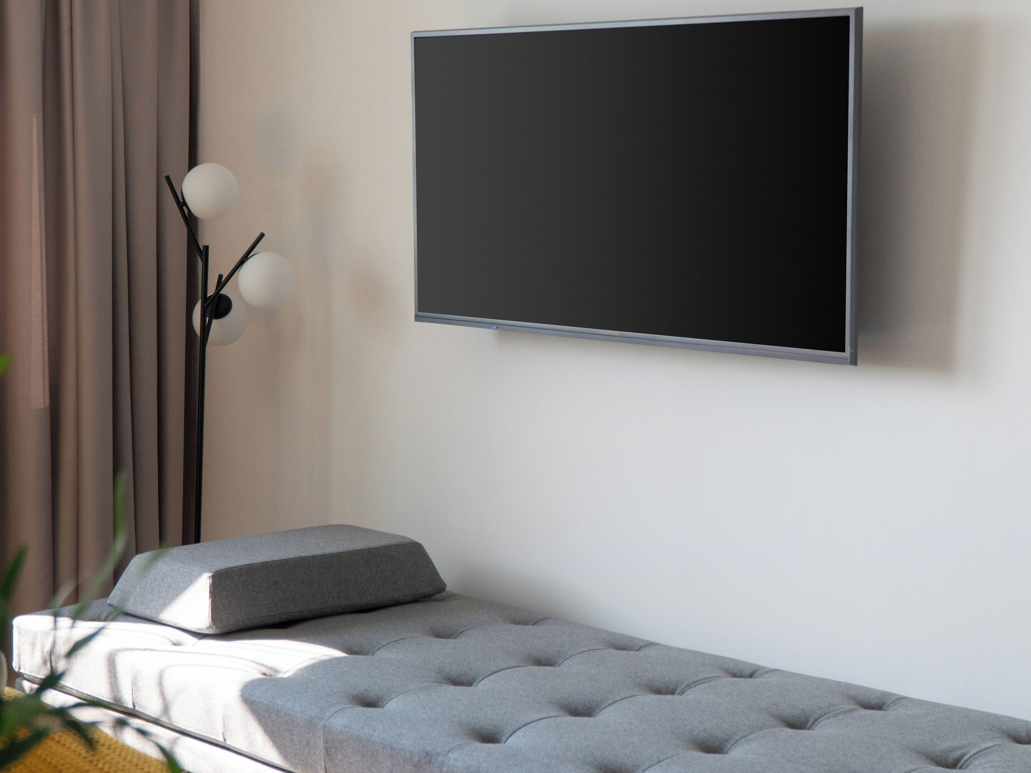 A modern room with a flat-screen TV mounted on the wall, a grey tufted bench, and a floor lamp with a spherical light.