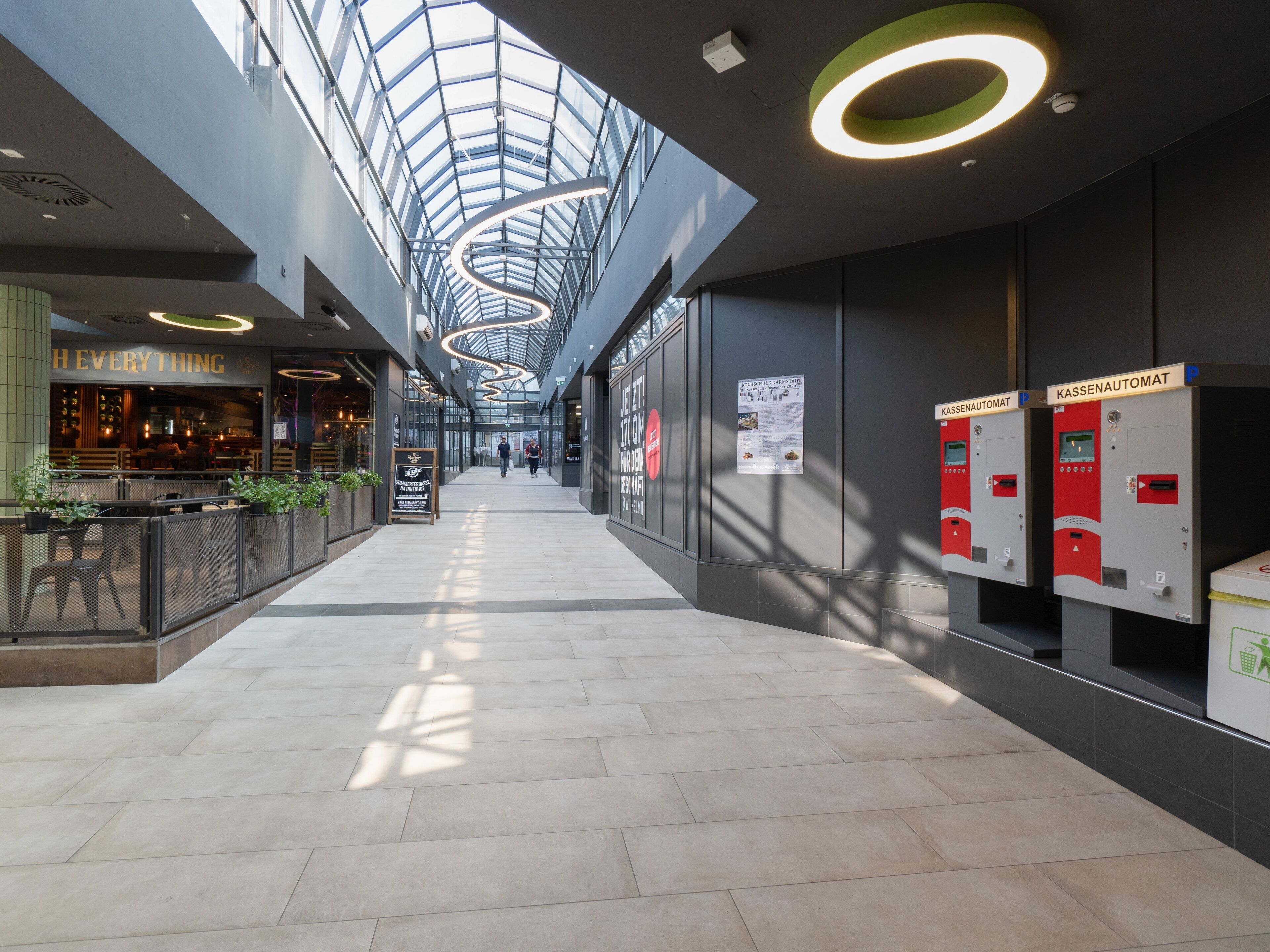 A modern indoor space with a high ceiling and skylights, featuring a restaurant, trash bins, and a clear walkway.