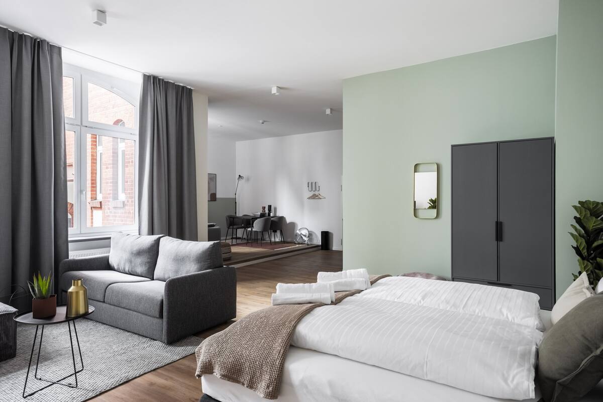 A modern hotel room with a grey sofa, a bed, a dining area in the background, and a large window with curtains.
