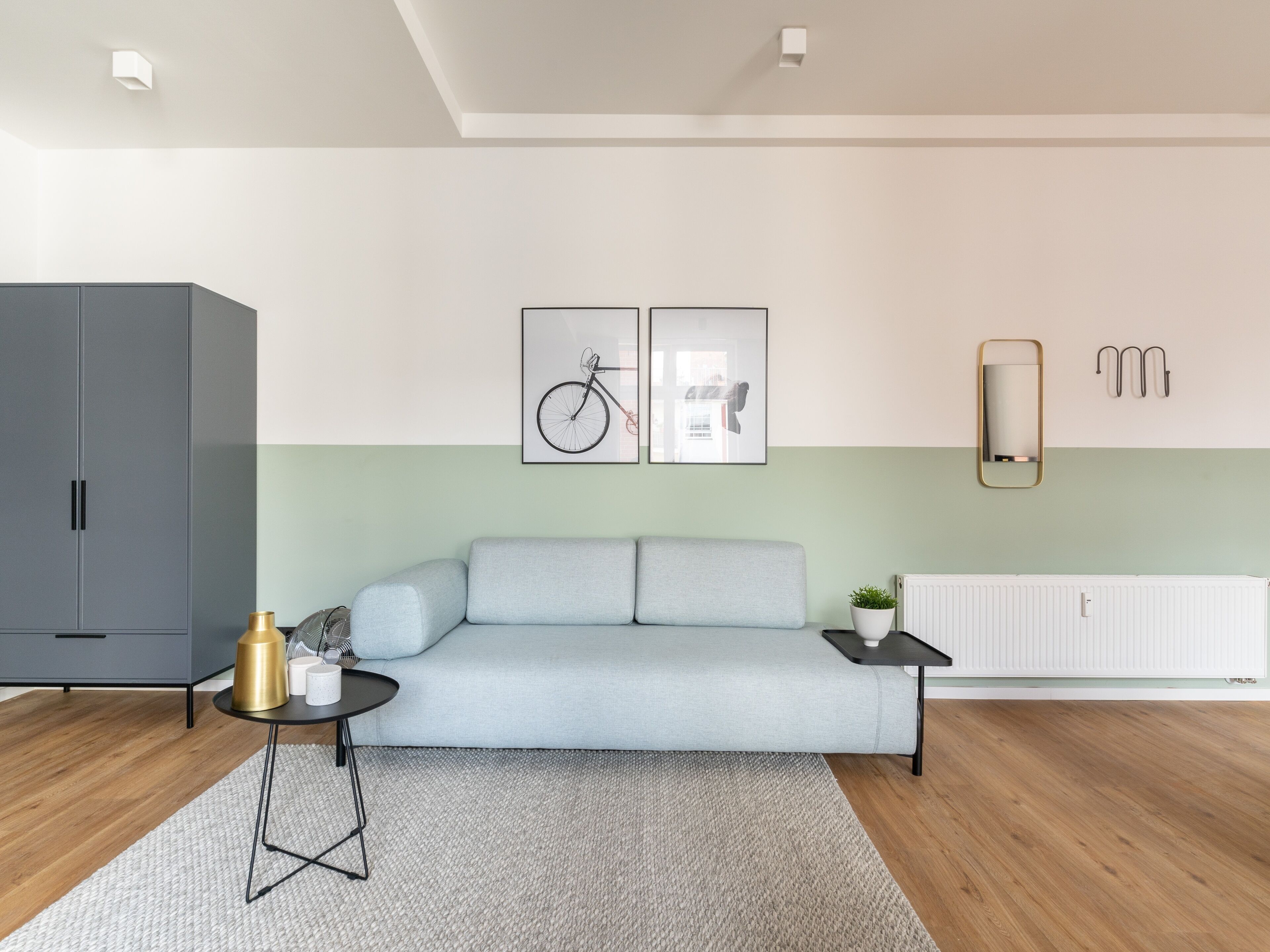 Una sala moderna con un sofá gris, una mesita redonda con una lámpara dorada y un radiador. Hay dos cuadros enmarcados en la pared y un espejo sobre el radiador.
