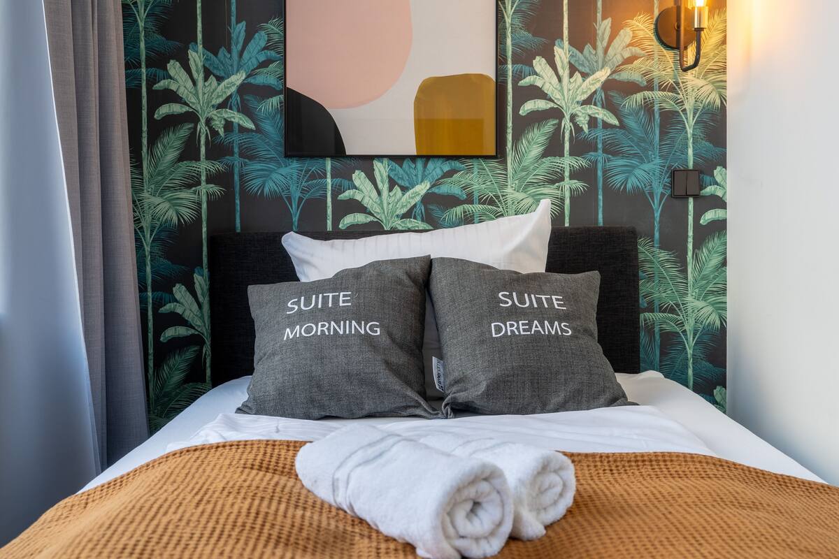A hotel room with a bed, two pillows labeled "SUITE MORNING" and "SUITE DREAMS," a wall with a tropical leaf pattern, and a wall-mounted light fixture.