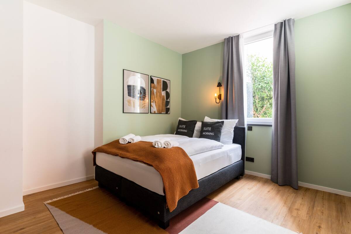 A neatly made bed with a brown blanket, a bedside table with a lamp, and a window with curtains.