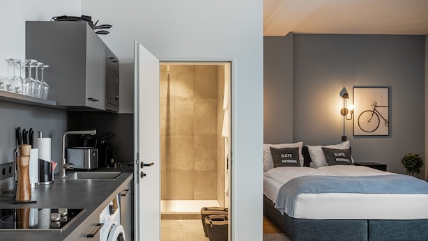 Una habitación de hotel moderna con kitchenette, una cama con almohadas a juego, una obra de arte de bicicleta colgada en la pared y un área de lavandería a la vista.