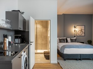 Une chambre d’hôtel moderne dotée d’une kitchenette, d’un lit avec des oreillers assortis, d’une œuvre d’art représentant un vélo accrochée au mur et d’un espace buanderie visible.