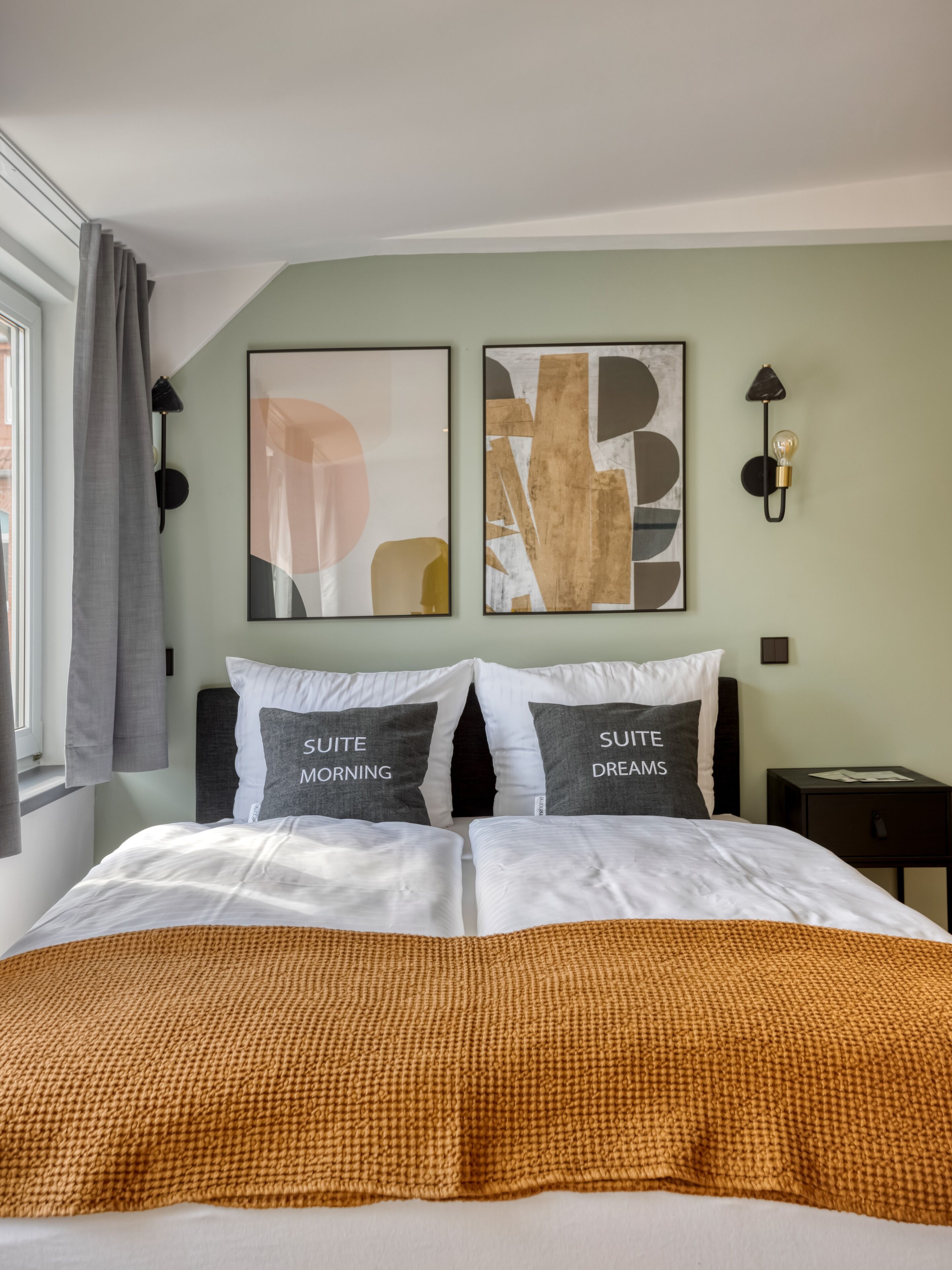 Une chambre à coucher avec un lit, deux oreillers ornés de motifs, une œuvre d’art abstraite accrochée au mur, une fenêtre avec des rideaux et une table de chevet.