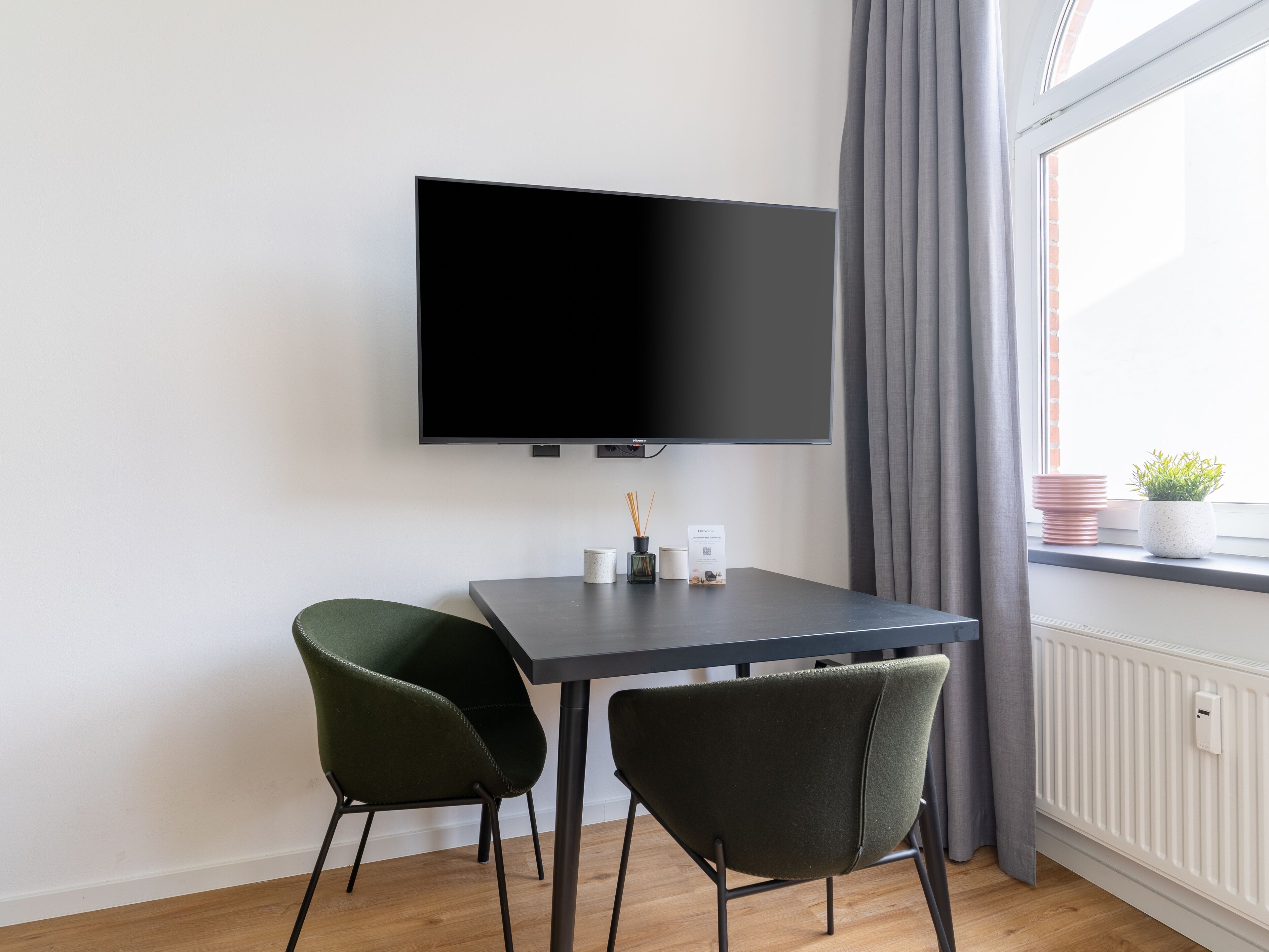 Habitació moderna amb televisió de pantalla plana penjada a la paret, una taula petita amb dues cadires, una finestra amb cortines i un radiador.