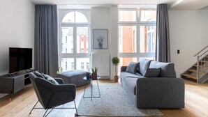 Una sala de estar moderna con un sofá gris, un sillón negro y un televisor sobre un mueble bajo. Se aprecia un ventanal con cortinas, una planta y una escalera a la derecha.