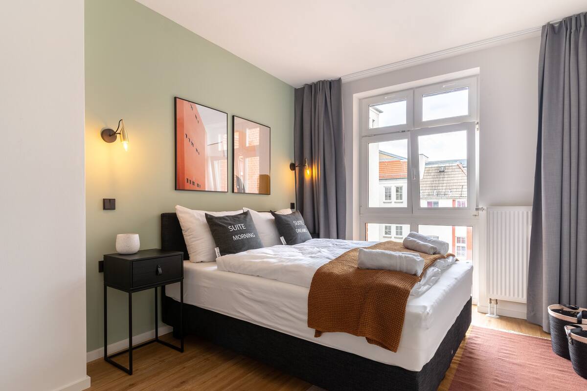 A modern bedroom with a large bed, a nightstand, a wall-mounted lamp, and a window with curtains.