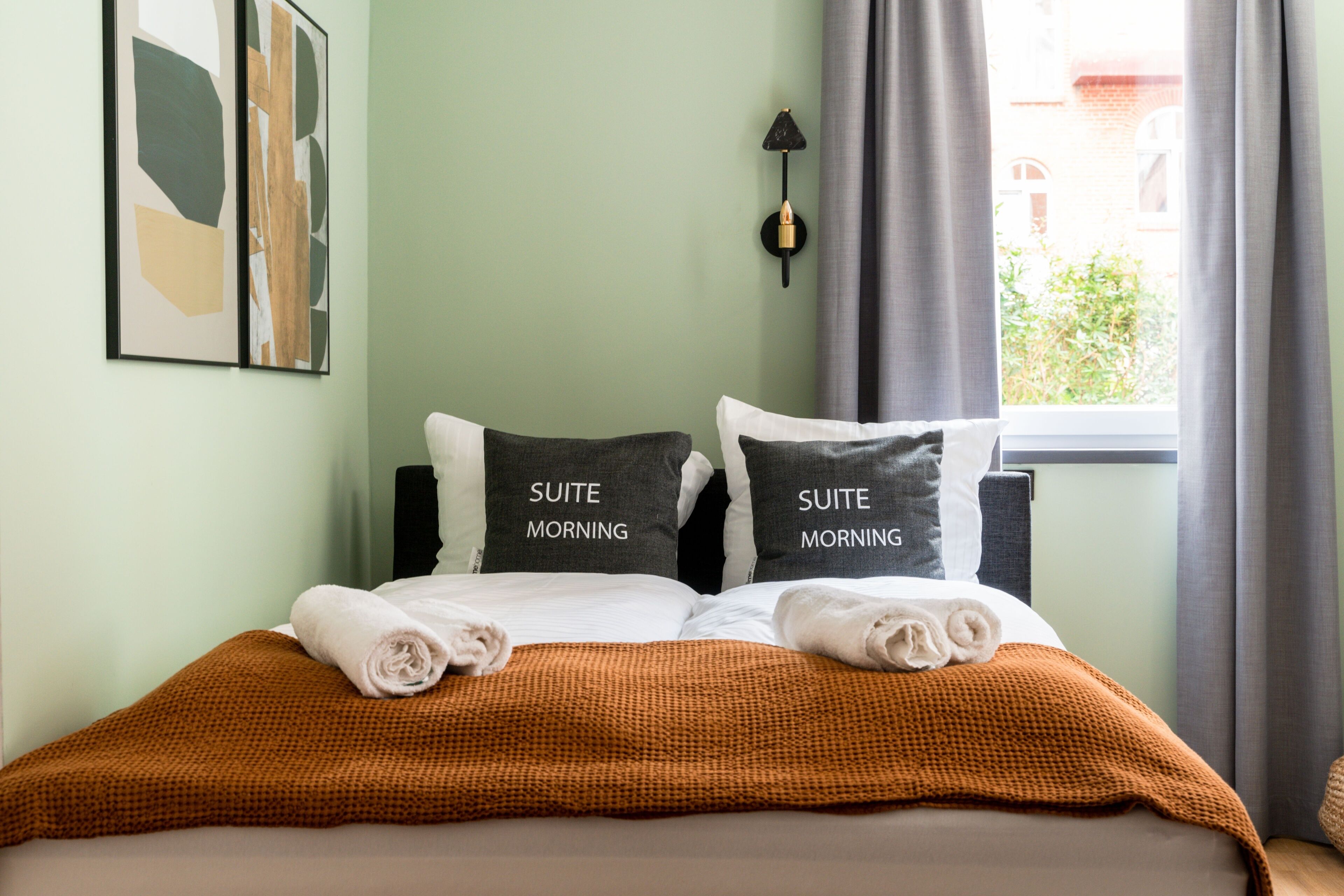 A neatly made bed with two pillows labeled "SUITE MORNING" and a brown checkered blanket.