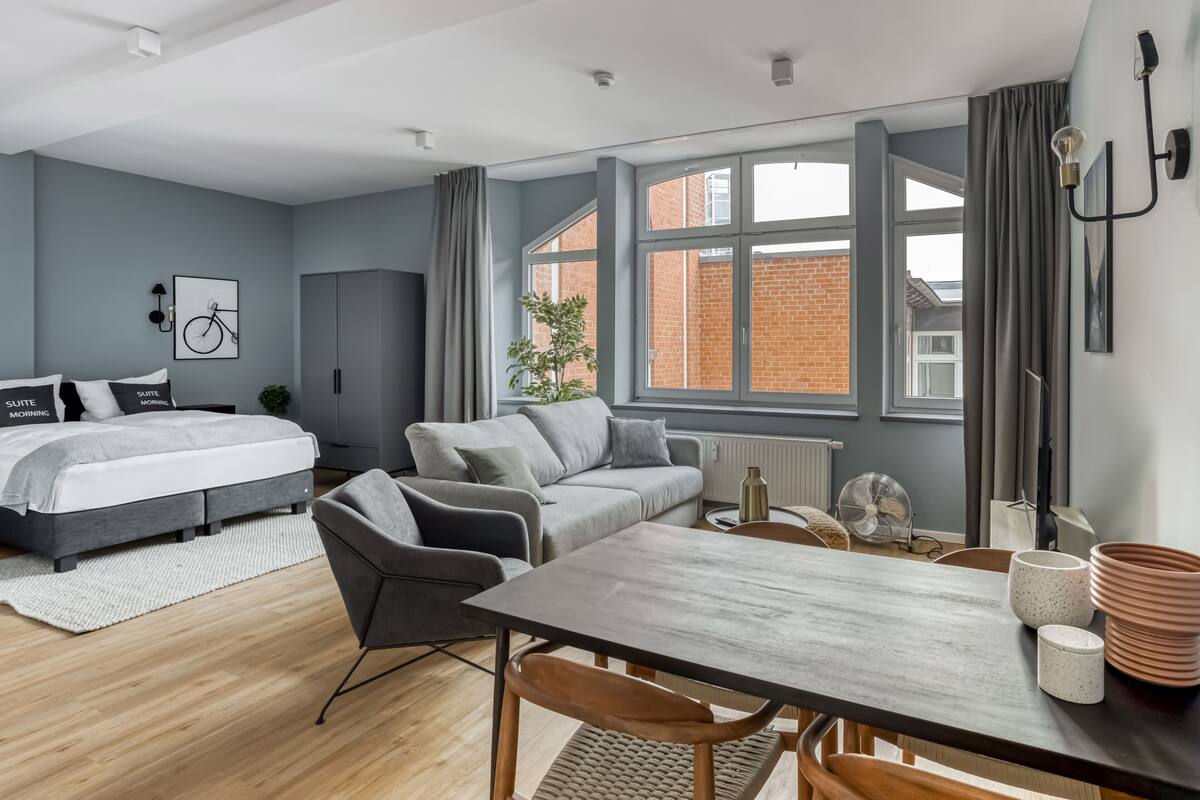 A modern bedroom with a bed, a grey sofa, a wooden dining table, and a large window with a view of a brick building.