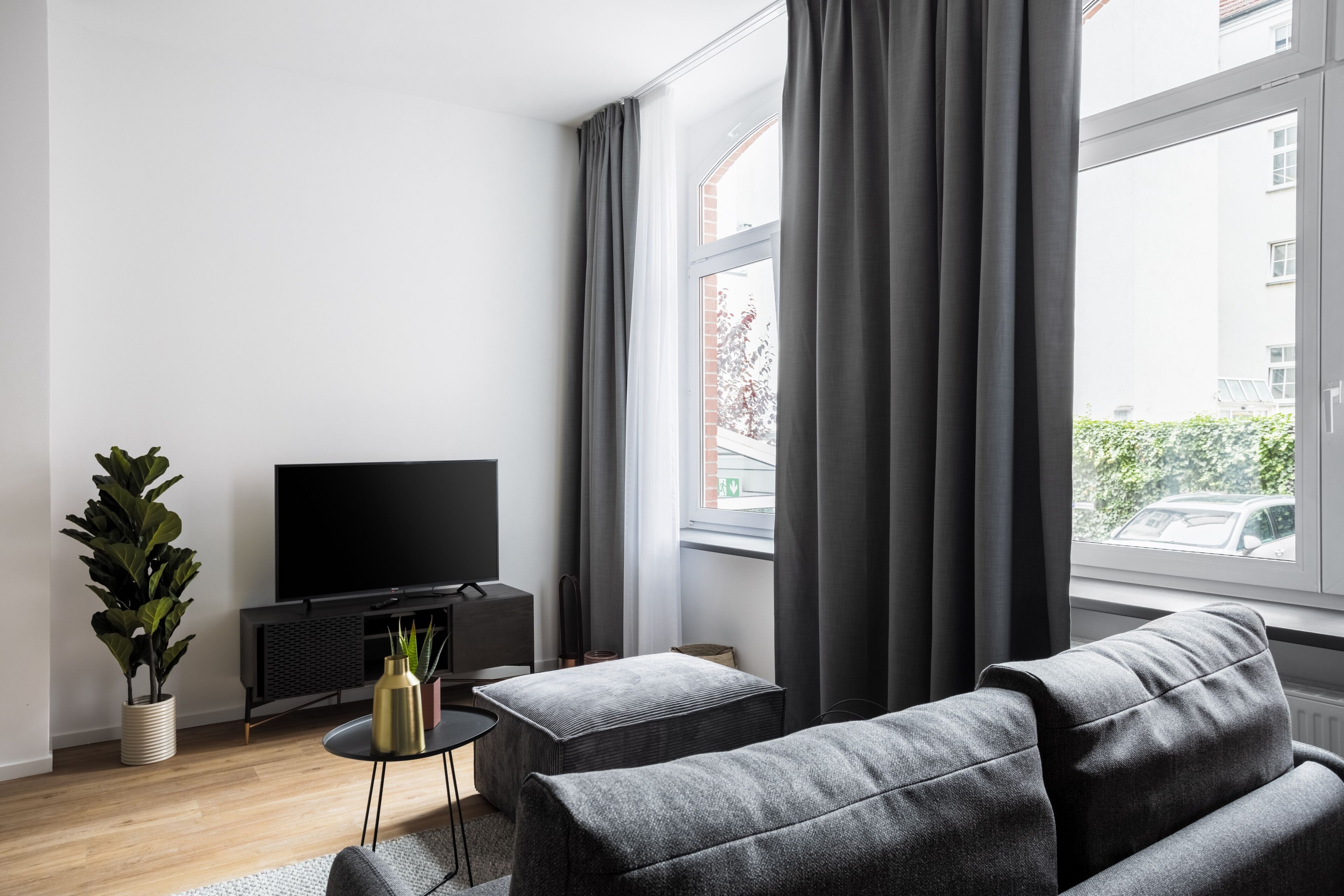A modern living room with a flat-screen TV, a grey sofa, a small side table, a potted plant, and a window with curtains.