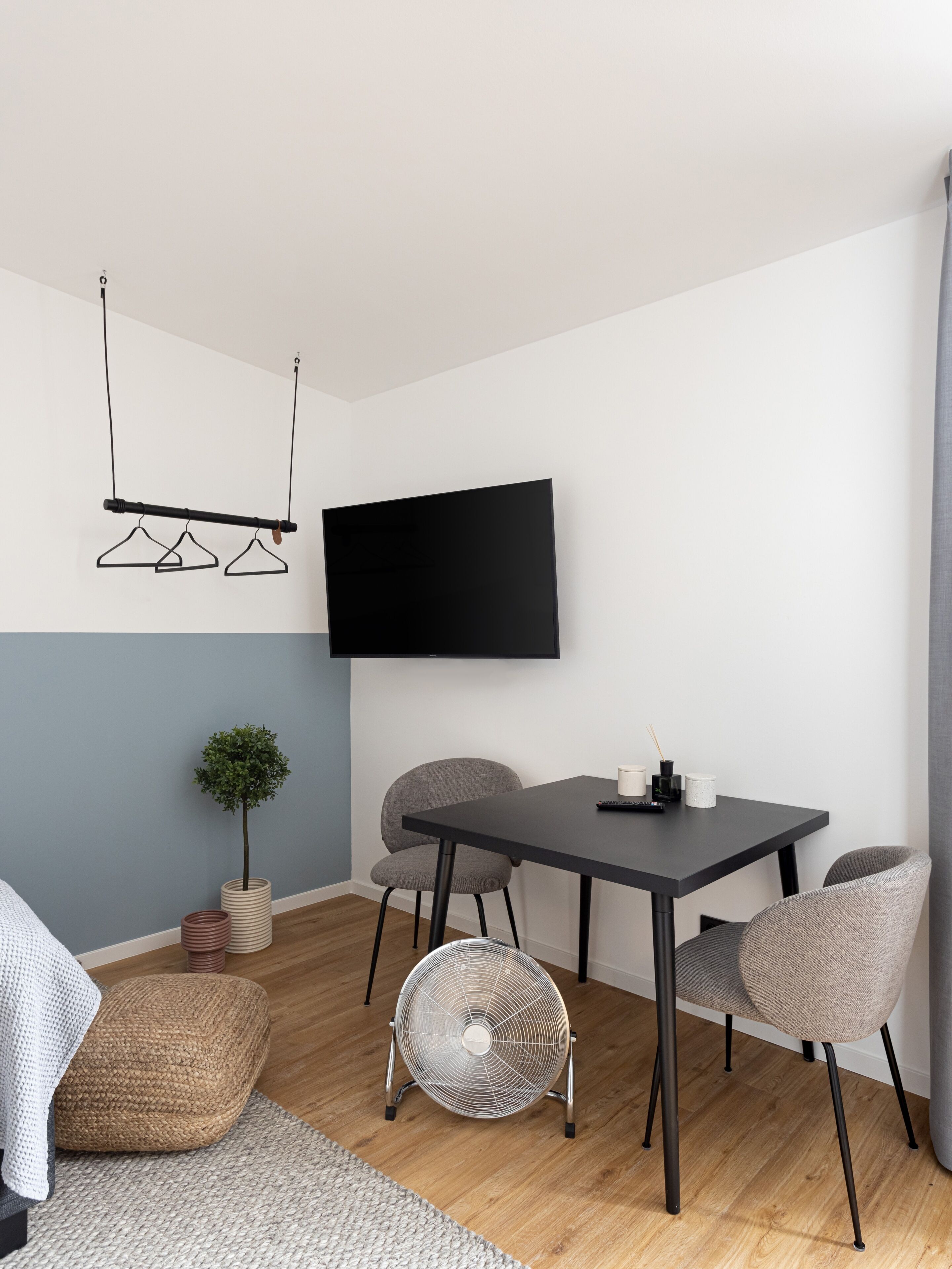 A modern living room with a flat-screen TV mounted on the wall, a dining table with chairs, a small potted plant, and a standing fan.