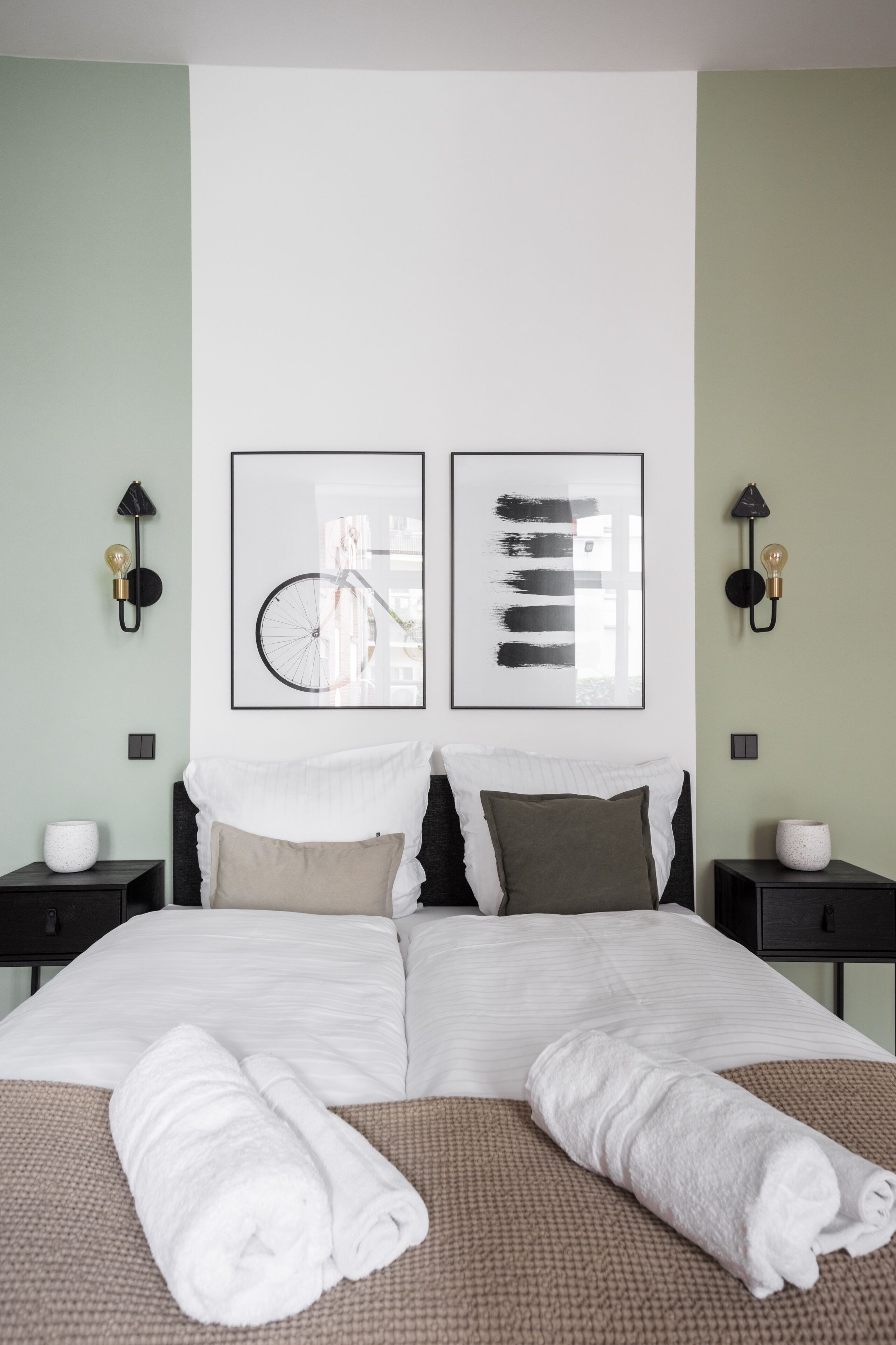 Una cama bien hecha con sábanas blancas y toallas enrolladas, un cuadro abstracto enmarcado y una lámpara de pared sobre una pared verde claro.