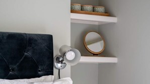Een keurig opgemaakt slaapkamer met een bed, kussens, een bedlampje, een ronde spiegel en planken met decoratieve objecten.