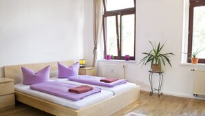 Cama doble con estructura de madera y colchón blanco, dos almohadas moradas y una colcha roja. Junto a la ventana hay una mesita con una planta en maceta.