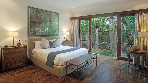 A bedroom with a large bed, wooden floor, a view of a garden through a glass door, and a painting on the wall.