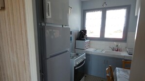 Eine kompakte Küche mit Kühlschrank, Herd und Mikrowelle. Über der Spüle befindet sich ein Fenster, im Vordergrund ist ein Stuhl zu sehen.