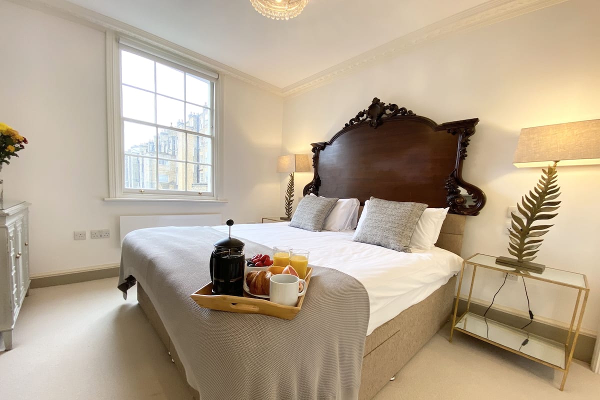 A neatly made bed with a tray of breakfast items, a large window, and a lamp on a side table.