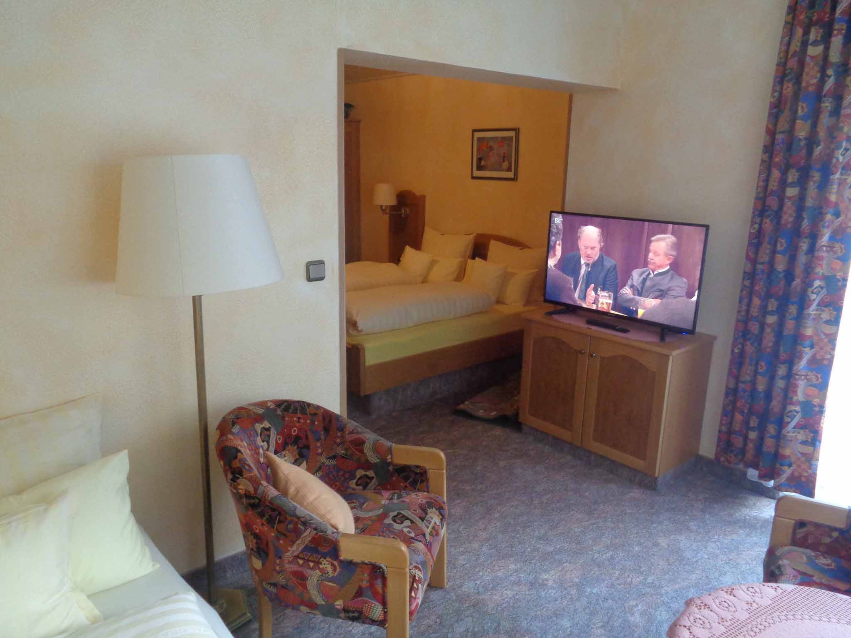 A hotel room with a bed, a television, a patterned armchair, and a small table.