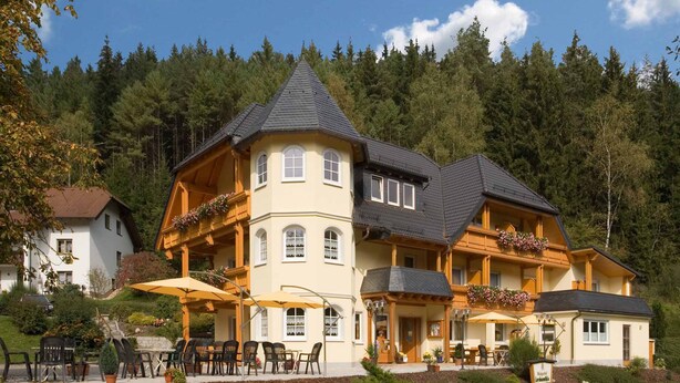 A large, multi-story building with a prominent balcony and a steeply pitched roof, surrounded by dense forest.