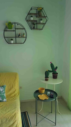 A room with a yellow sofa, a round coffee table with a phone and a bowl, a small white table with two potted cacti, and two wall-mounted shelves with plants and figurines. - 28 · Appartamento sul mare (Marina Julia)