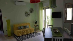 A living room with a yellow sofa, a dining table with chairs, a small side table, and a TV mounted on the wall. - 28 · Appartamento sul mare (Marina Julia)