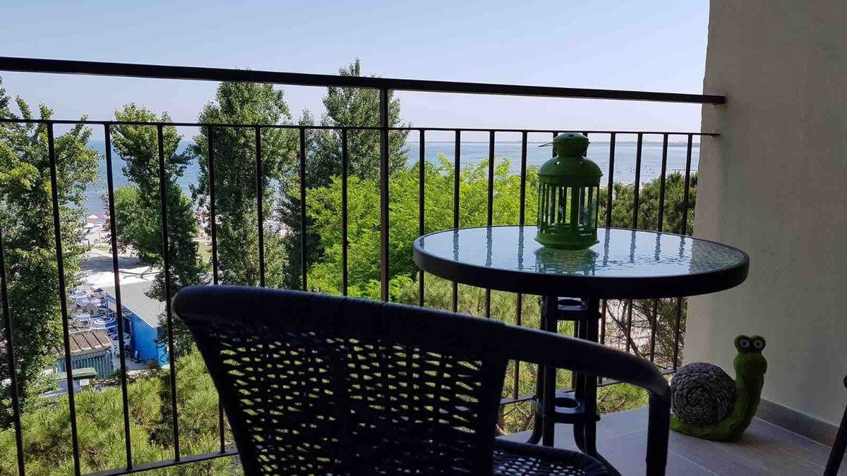 Een balkon met een ronde tafel en een stoel, met uitzicht op bomen en een wateroppervlak.