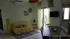 Uma sala de estar com um sofá amarelo, uma mesa de jantar com cadeiras, uma TV fixada na parede e uma janela com cortinas.