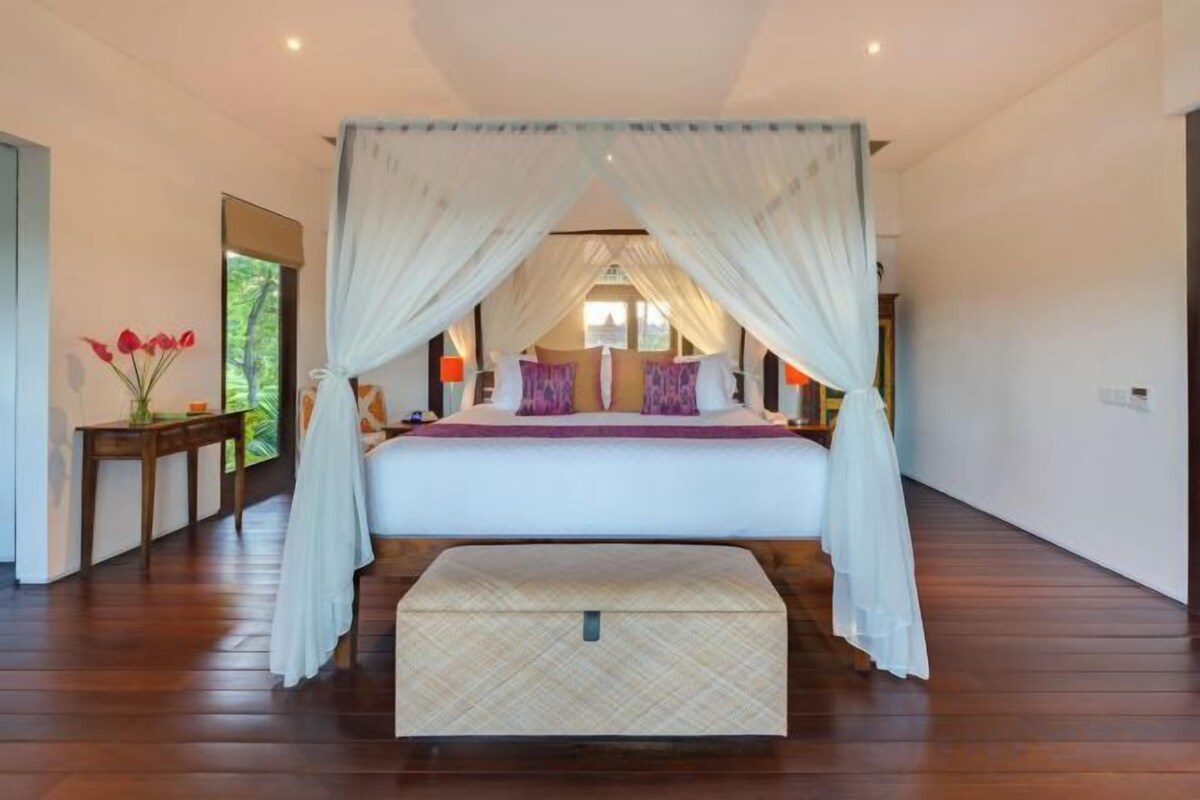 A bedroom with a canopy bed, bedside tables, and a view of greenery outside.
