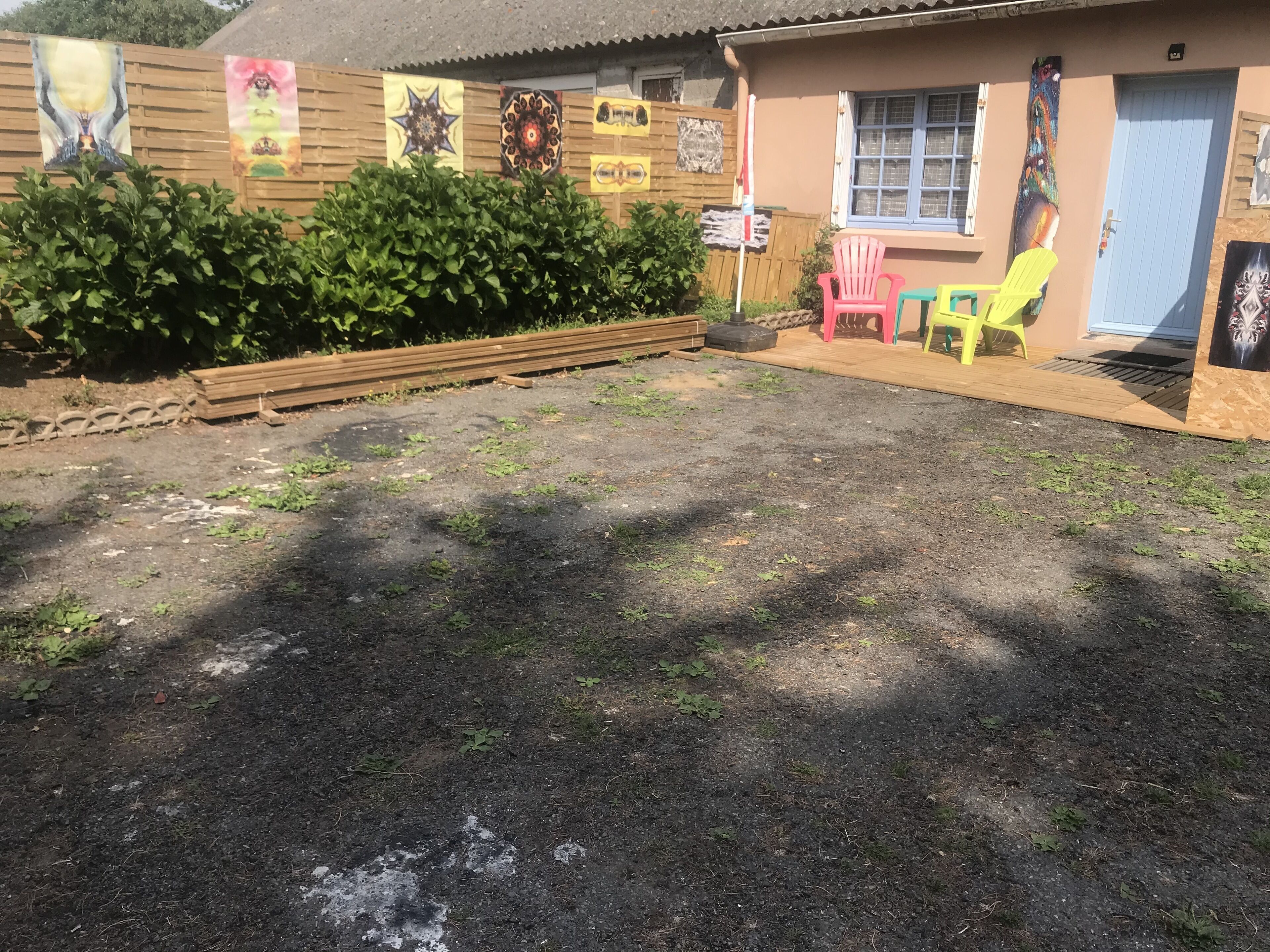 Un edificio con cercado de madera, sillas coloridas y un área pavimentada con césped escaso.